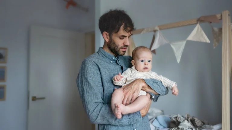 Man in a blue shirt holding a baby dressed in a striped onesie in a nursery.