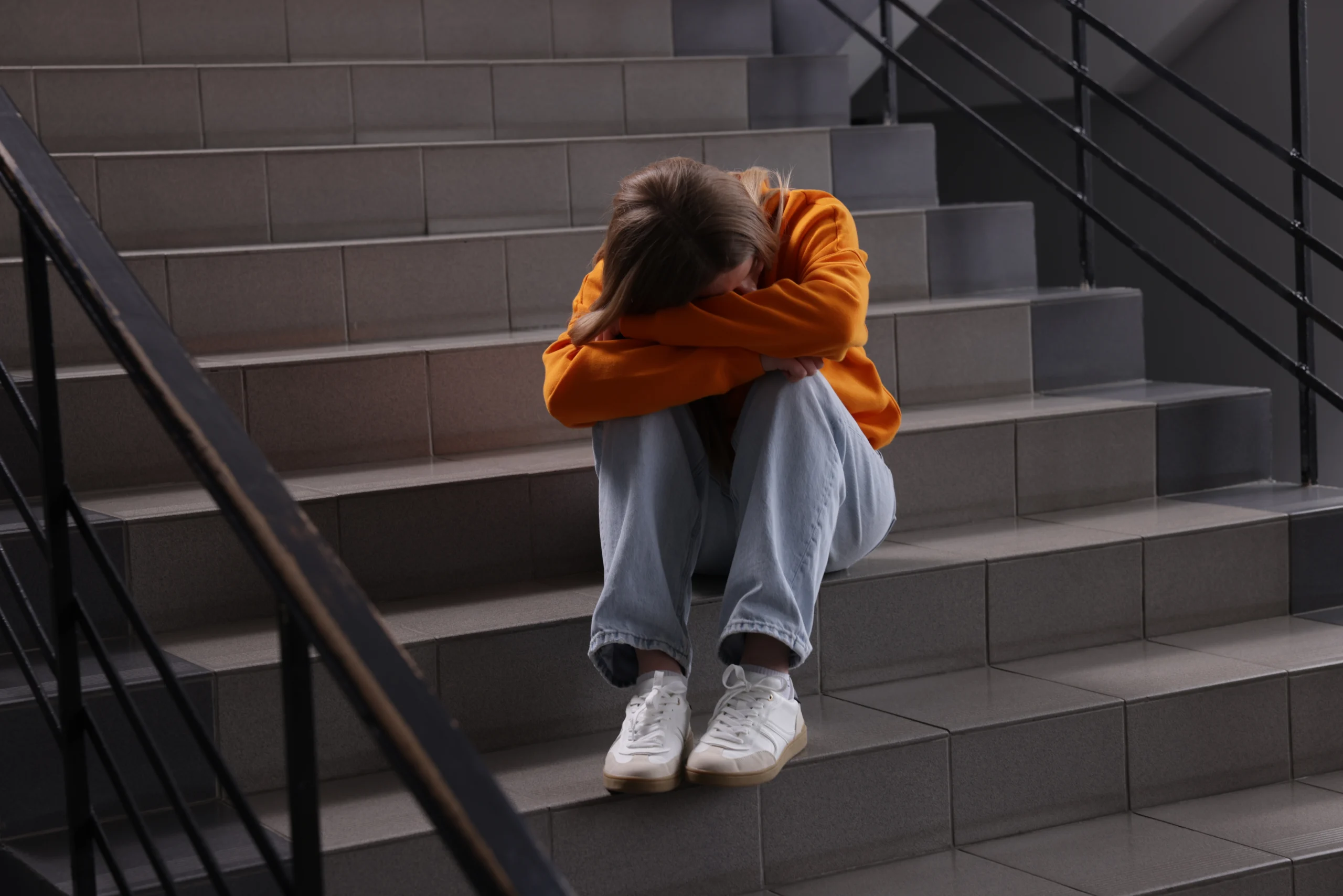Young woman in orange sweatshirt sitting on gray tiled stairs with head resting on folded arms.