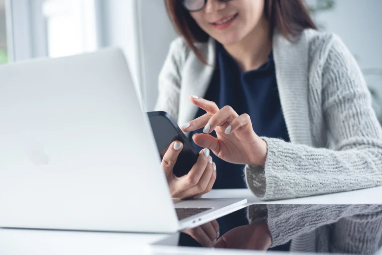 Woman wearing glasses and a gray sweater using smartphone and laptop at desk.