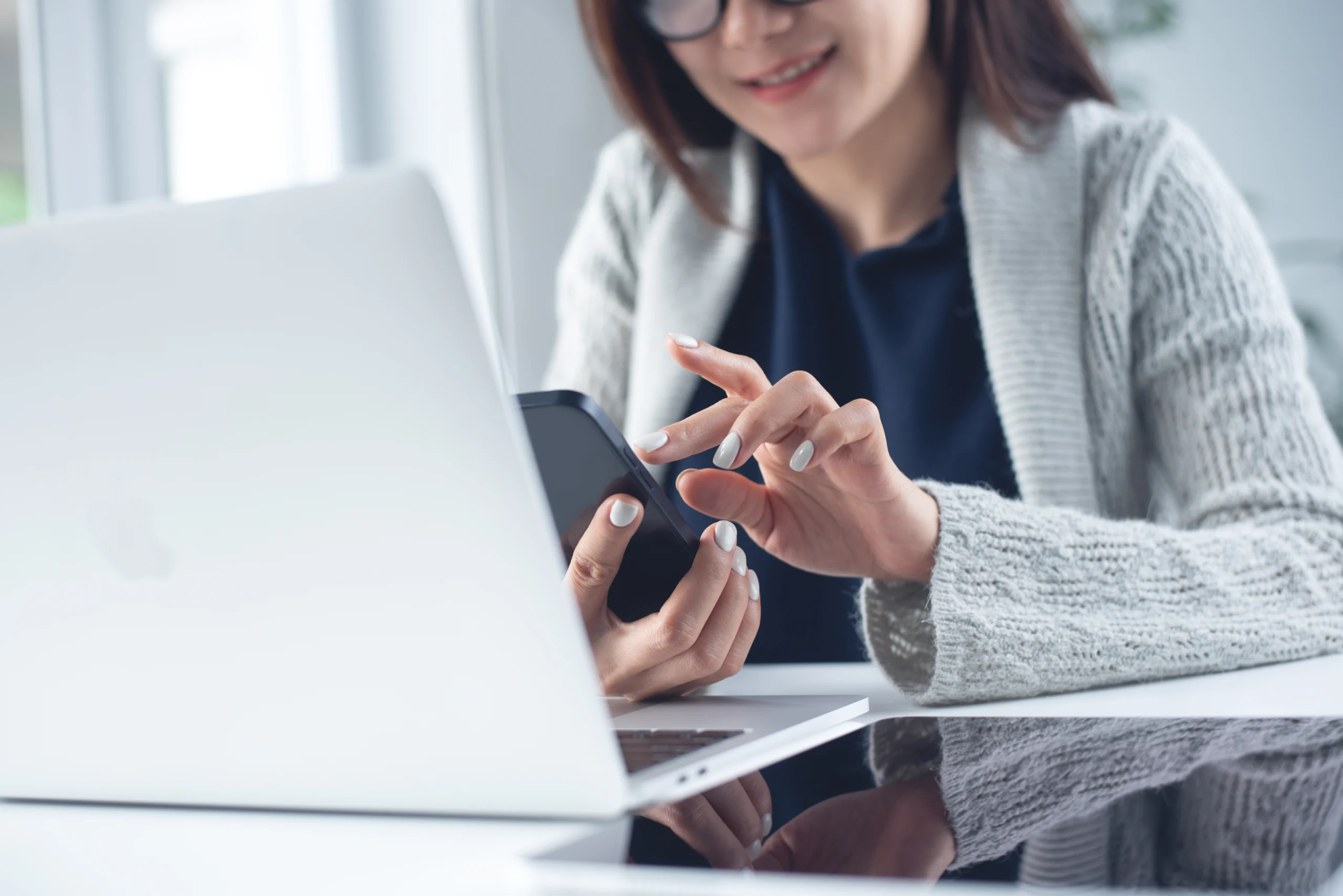 Woman wearing glasses and a gray sweater using smartphone and laptop at desk.