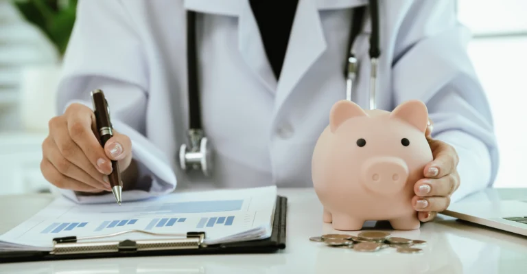 Medical professional in white coat holding piggy bank and pen over financial documents.