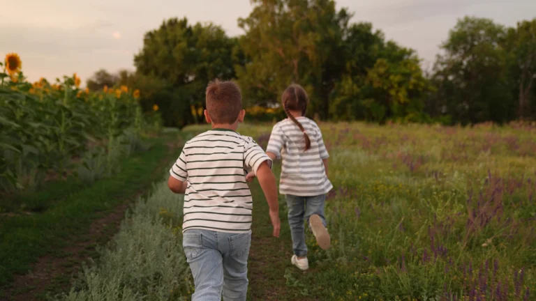 Two children in striped shirts running on a grassy path near sunflower field at sunset.