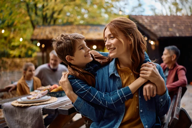 Mother and son embracing outdoors with family gathering and autumn foliage background.
