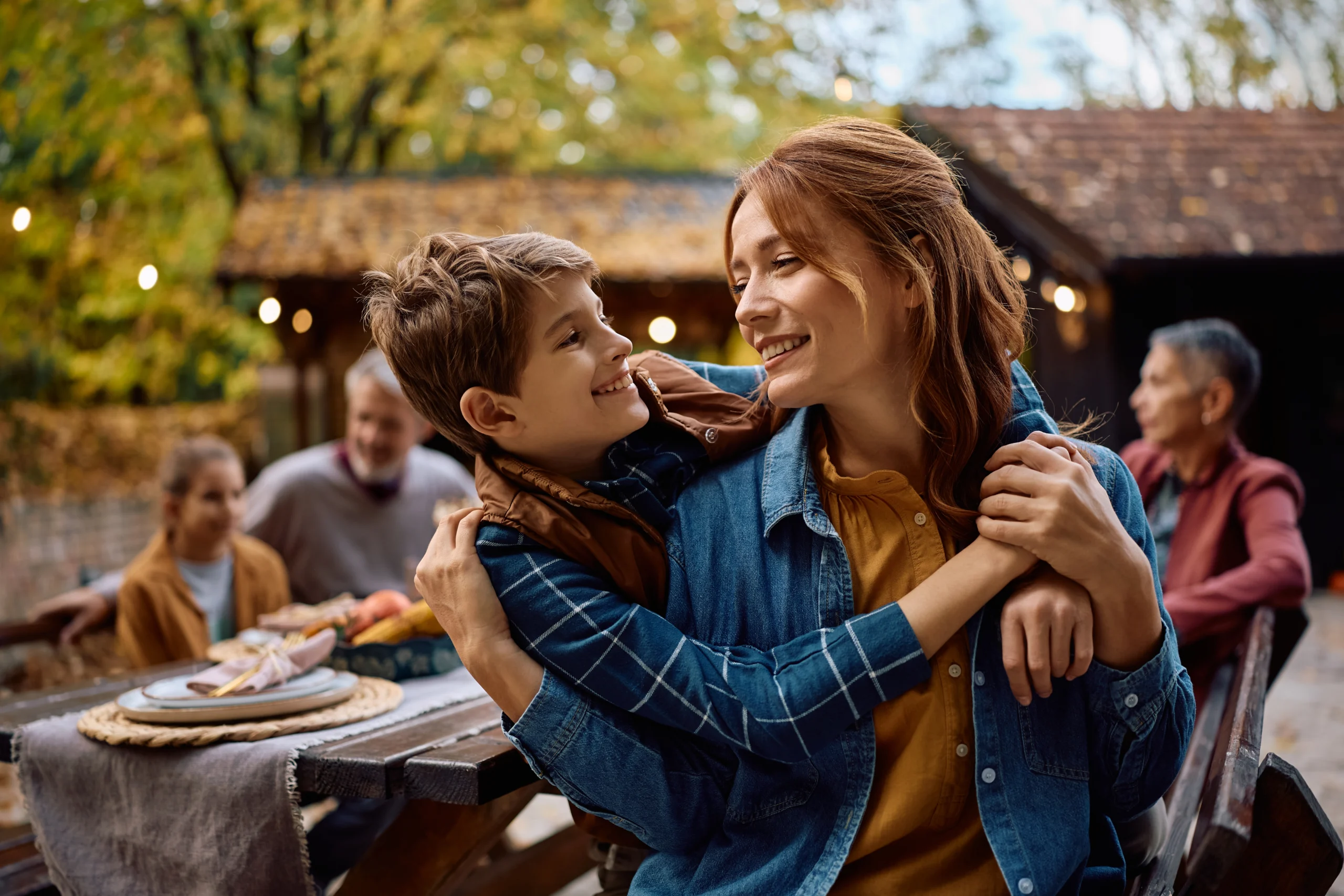 Mother and son embracing outdoors with family gathering and autumn foliage background.