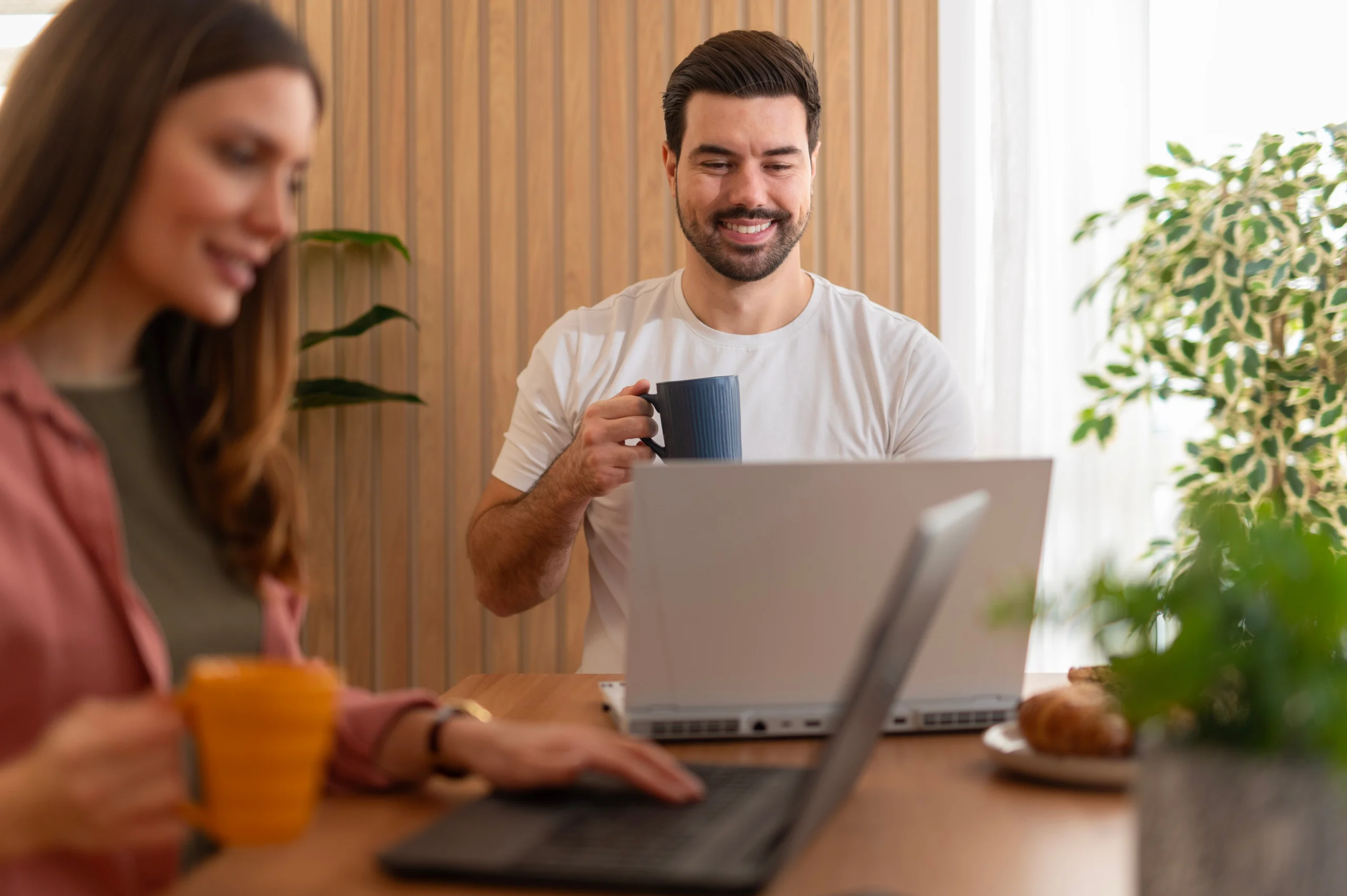Two professionals working on laptops in a modern office with wooden paneling and plants.