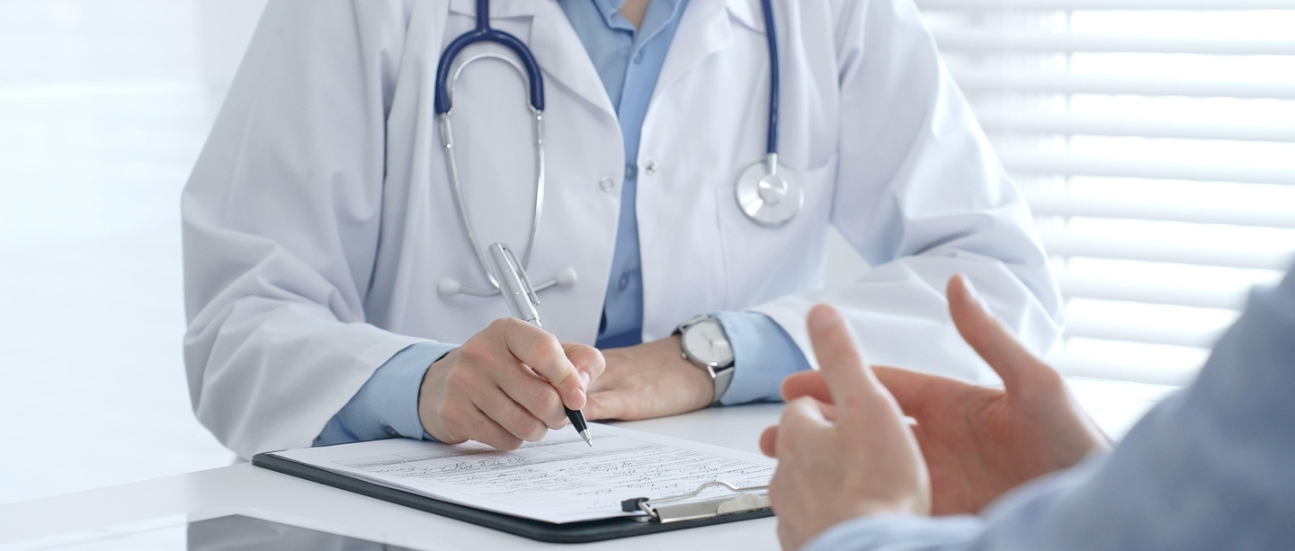 Doctor in white coat with stethoscope writing on clipboard during patient consultation.