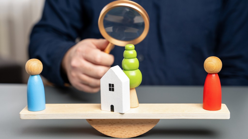 Person holding magnifying glass over wooden balance scale with house, tree, and two figurines.