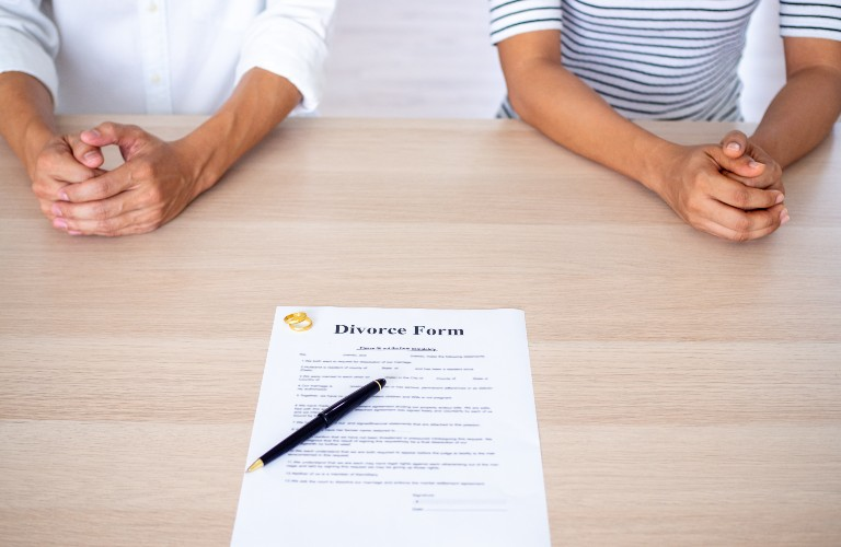 Two individuals sitting apart at a table with a divorce form and pen placed between them.