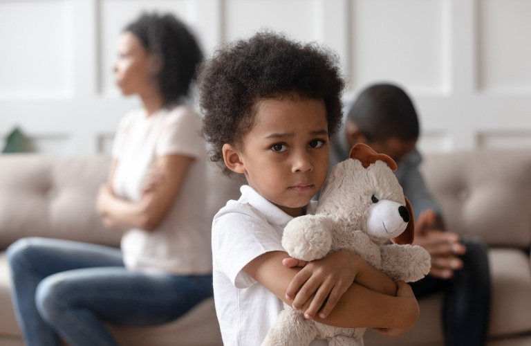 Young boy clutching a stuffed dog, with two adults sitting apart in the background.