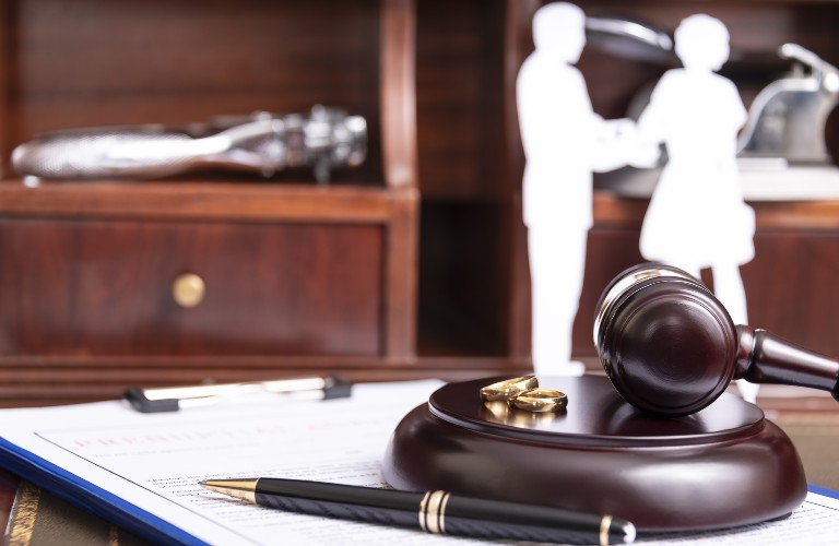 Wooden judge's gavel with gold wedding rings on base, pen, and paper, blurred couple cutouts.