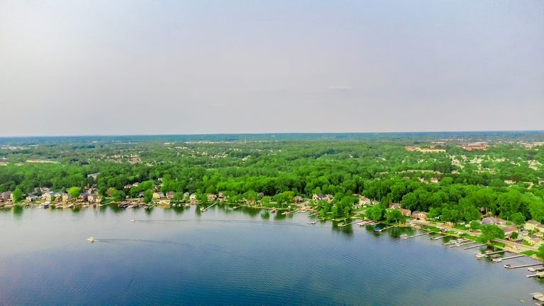 Aerial view of a lake with surrounding residential homes and dense green trees under a clear sky.
