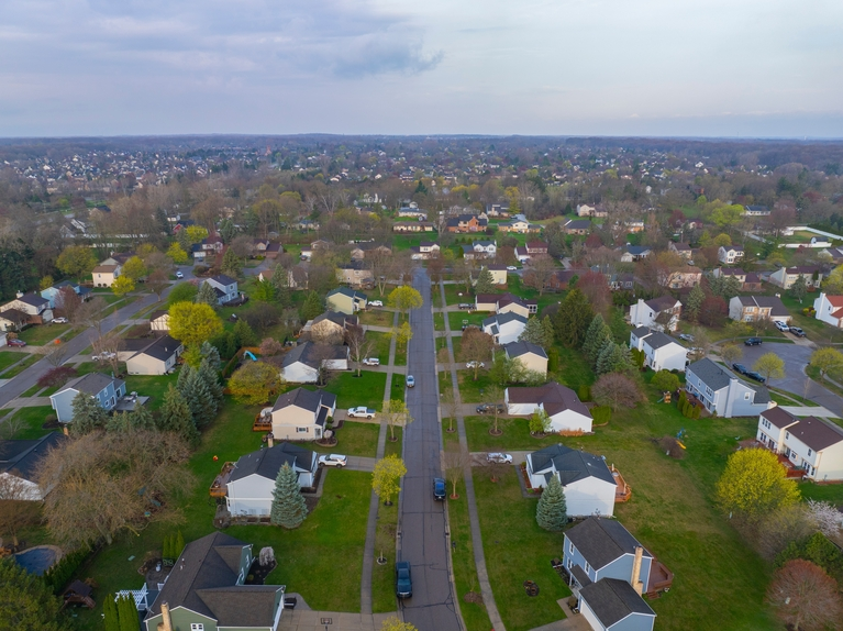 Aerial view of a suburban neighborhood with houses, green lawns, trees, and a central street under a cloudy sky.