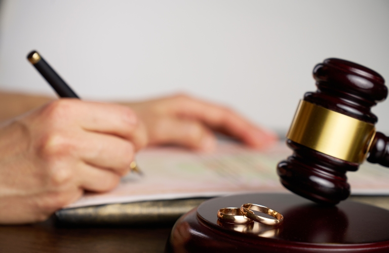 Close-up of a wooden judge's gavel with two gold wedding rings on a desk, blurred hand writing in the background.