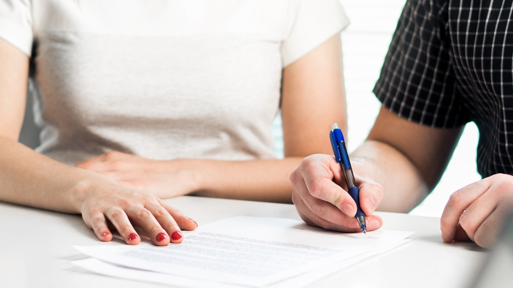 Two people at a table, one signing a document with a blue pen, the other resting hands nearby.