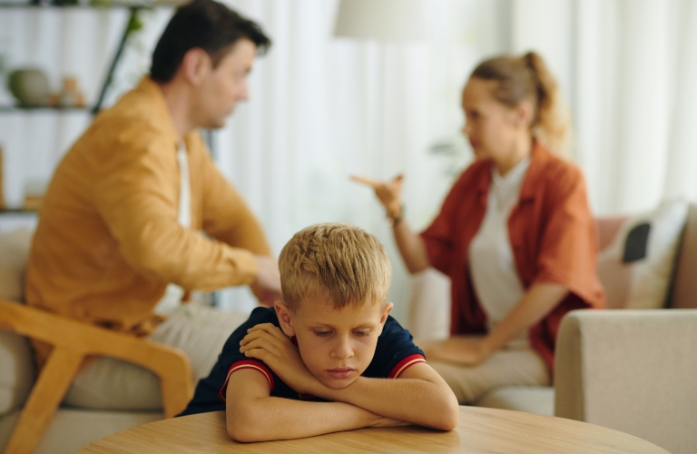 Young boy resting head on folded arms at table, parents arguing in background.