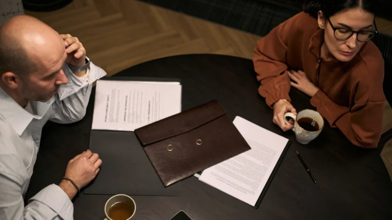 Two individuals seated at a dark round table with documents, coffee cups, and wedding rings.