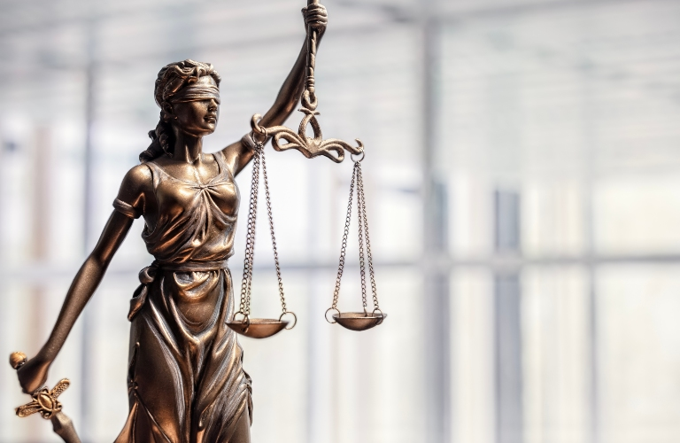 Bronze statue of Lady Justice blindfolded, holding scales and a sword, with a blurred modern office background.