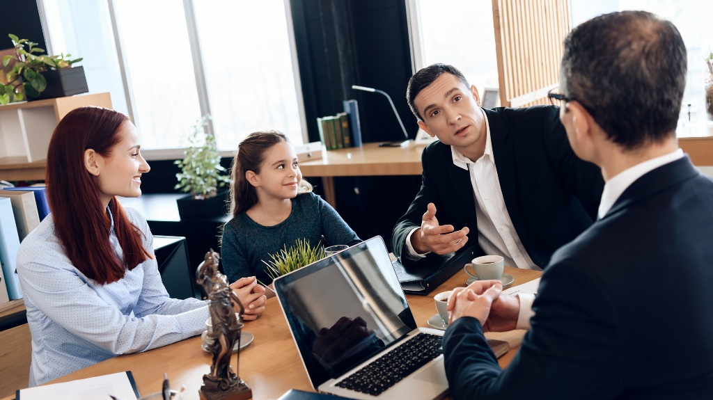 Four people, including a young girl, engaged in a discussion around a wooden office table with a laptop and coffee cups.