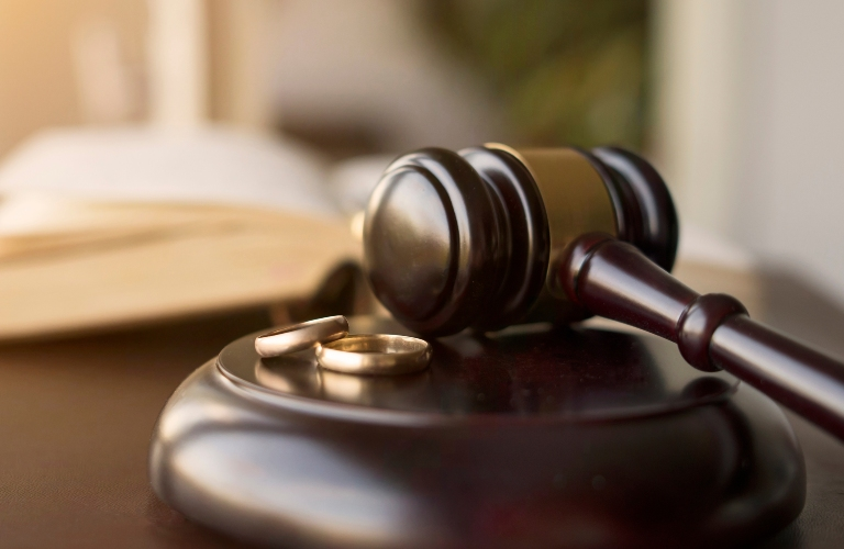 Dark wooden judge's gavel resting on a block with two gold wedding rings beside it.
