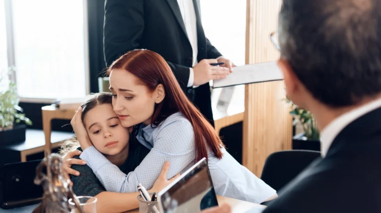 Woman comforting a young girl in an office setting while two men discuss documents nearby.