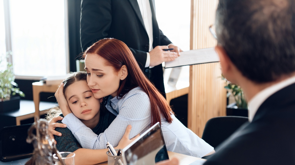 Woman comforting a young girl in an office setting while two men discuss documents nearby.